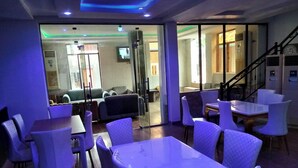 A modern hotel lobby with a seating area featuring white tables and chairs, a staircase, and a view into the adjacent living area. - KINGSAMYSTAR HOTEL AND SUITES  (Asaba)