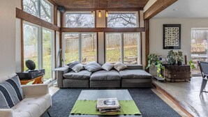 A cozy living room with a grey sofa, a white armchair, and a coffee table with books. Large windows offer a view of the backyard. - Modern 5BR Home with Mtn Views, BBQ & Pool Table (Asheville)