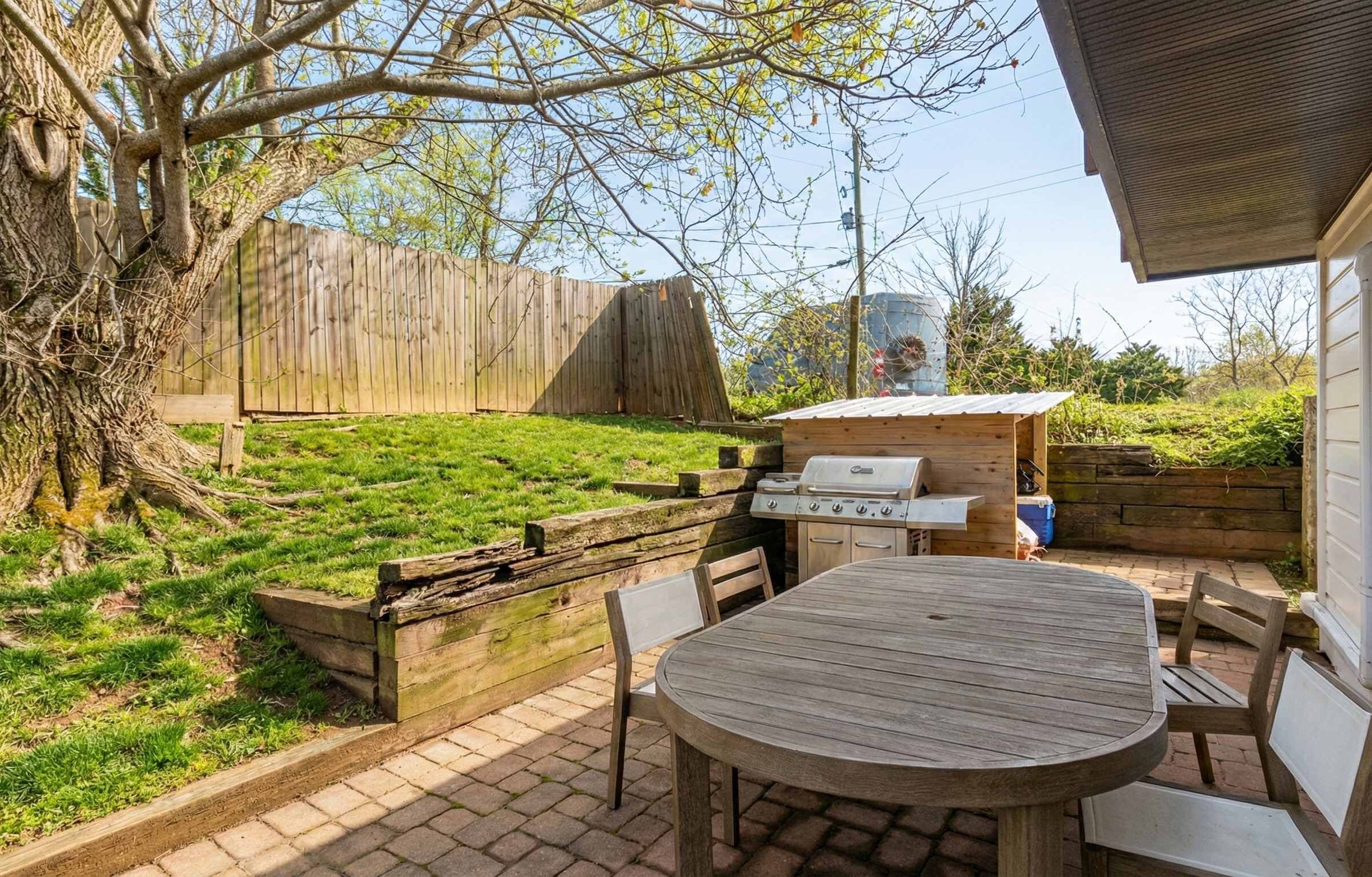 An outdoor patio with a wooden table and chairs, a grill, and a wooden fence.