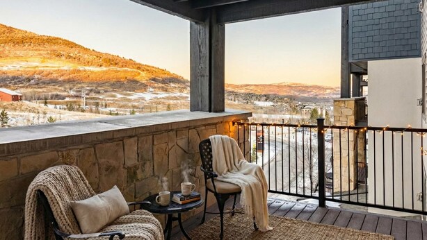 A balcony with a stone wall, a chair, a table with a mug and books, and a view of a valley with a building in the distance. - Silverado Suit - Steps to Canyons, Golf & Trails (Park City)