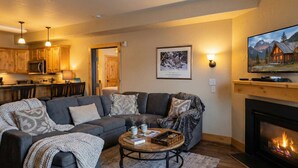A cozy living room with a sectional sofa, a coffee table with books and mugs, a fireplace, and a kitchen in the background. - Silverado Suit - Steps to Canyons, Golf & Trails (Park City)