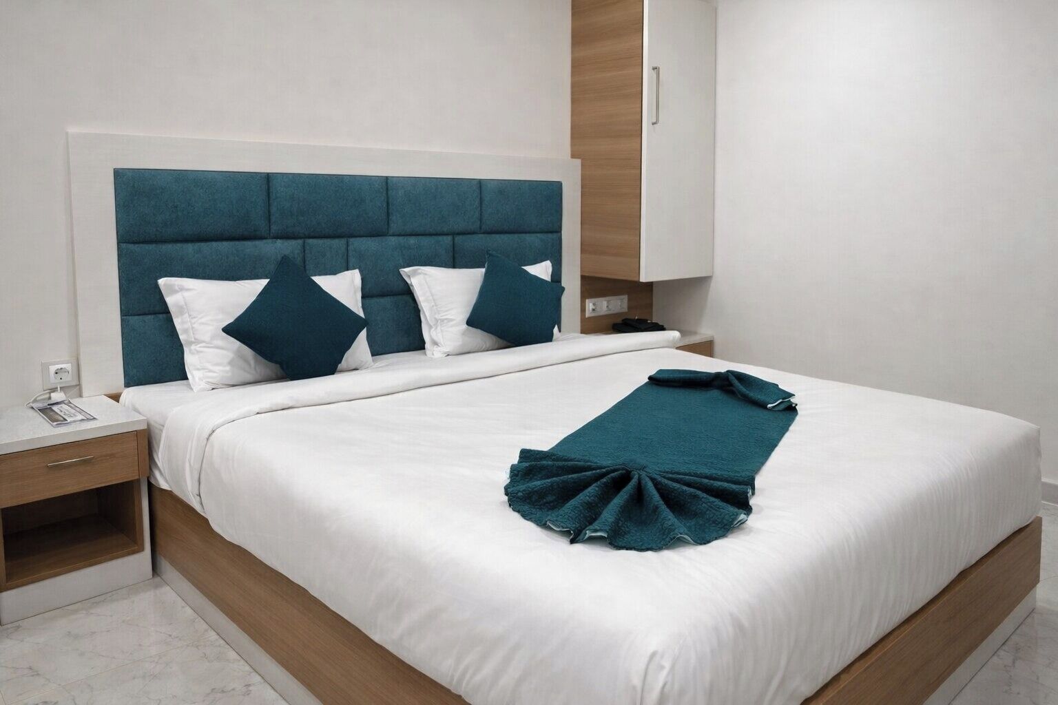 Un lit bien fait, avec une literie blanche et des coussins turquoise, une table de chevet en bois avec un téléphone et un magazine, et une tête de lit en bois.