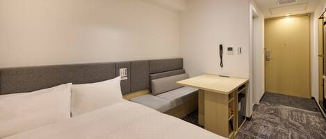 A hotel room with a bed, a small desk, a bench, and a door.