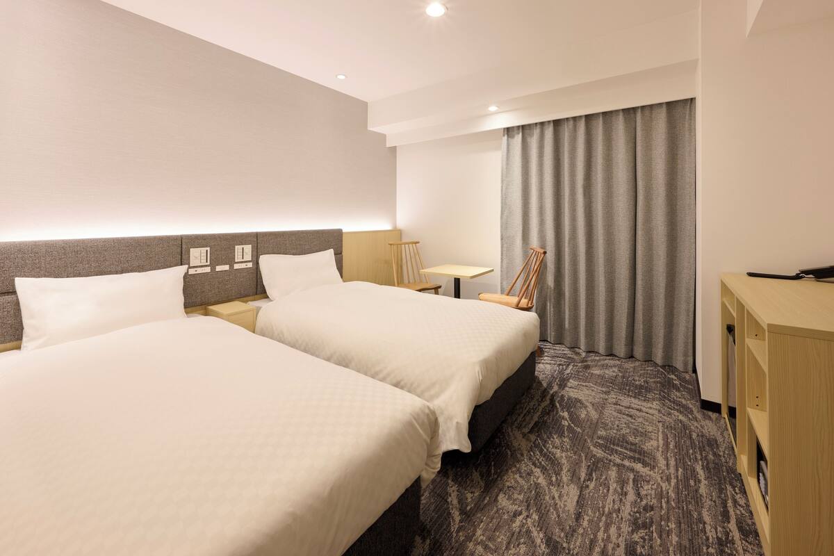 A hotel room with two beds, a desk, a chair, and a large window with curtains.