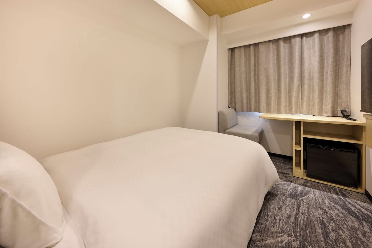 A hotel room with a bed, a desk, a chair, a small refrigerator, and a window with curtains.