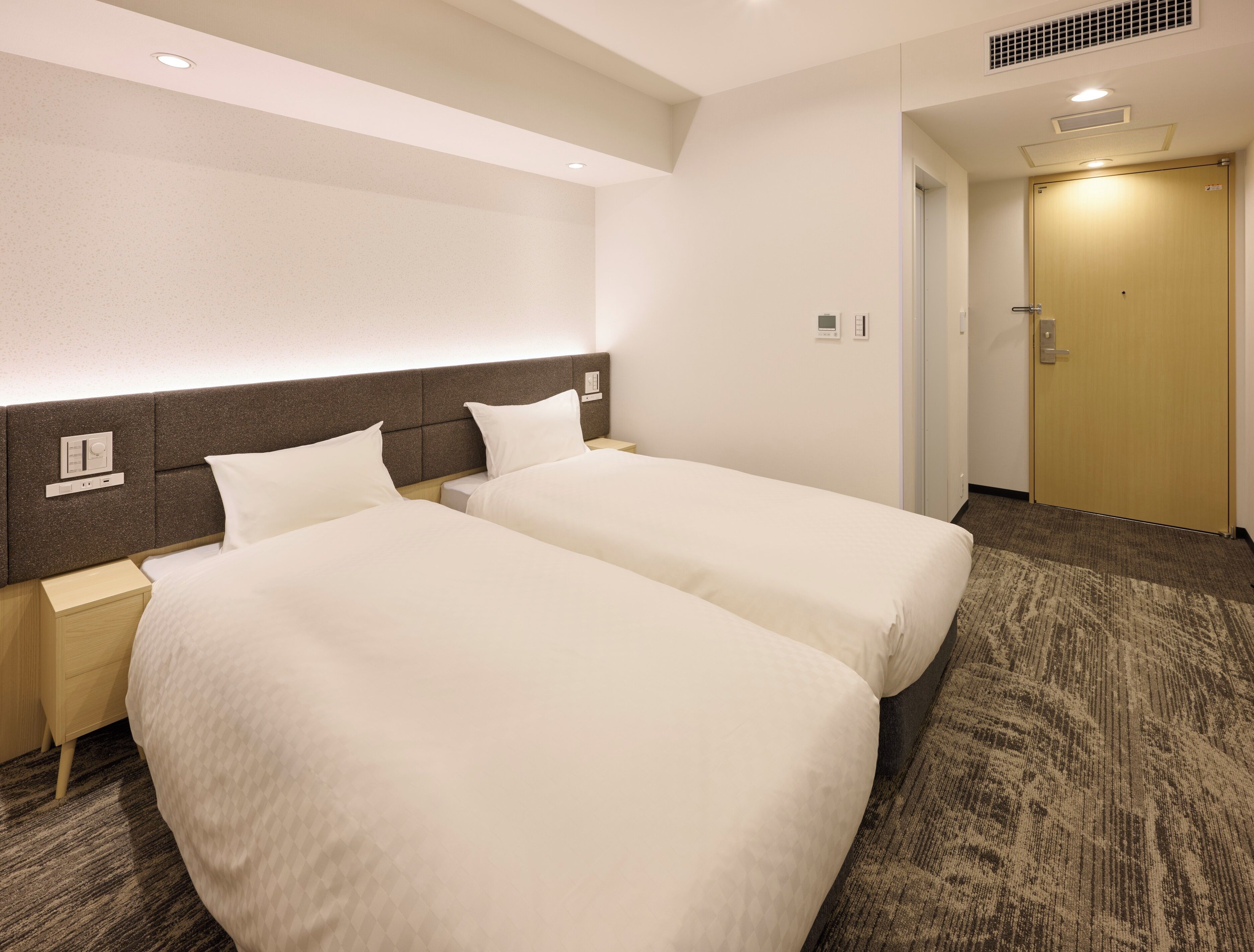 A hotel room with two beds, a nightstand, a wall-mounted control panel, and a door.