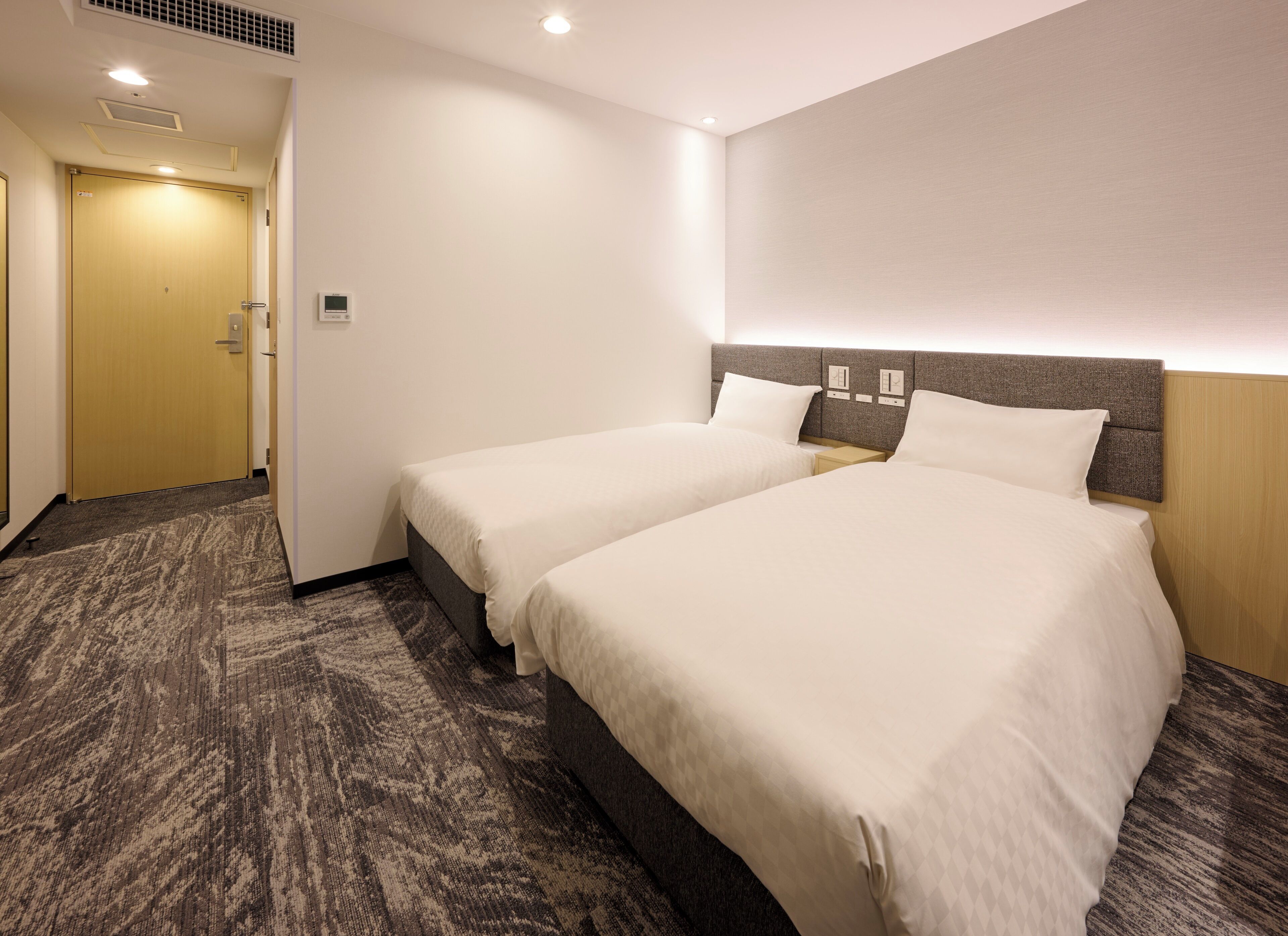 Une chambre d’hôtel avec deux lits, un sol recouvert de moquette, une porte en bois et un interrupteur sur le mur.