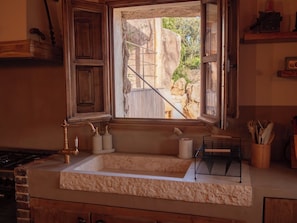 A kitchen with a stone countertop, a sink, a window with a view of a stone wall and greenery, and wooden cabinets. - Charming sheepfold in Corsica, heated pool, between scrubland and beautiful beaches. (Figari)