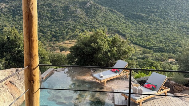 A wooden deck with a glass-bottomed pool, lounge chairs, and a view of a mountainous landscape.