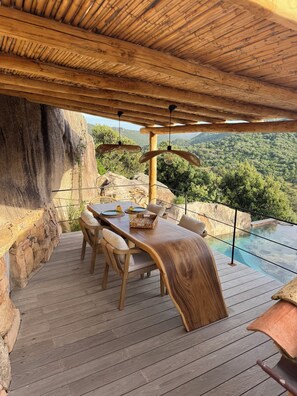 Una terraza de madera con una mesa y sillas de comedor, con vista a una piscina cristalina y abundante vegetación.