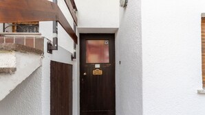 Ein schmaler, weiß getünchter Gang, der zu einer dunklen Holztür mit kleinem Fenster und einem Schild führt.