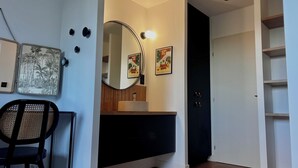 Une pièce avec un miroir rond, un meuble noir, une chaise et des œuvres d’art accrochées au mur.