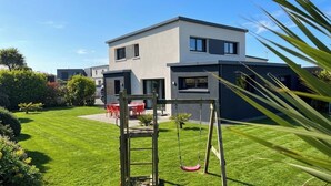 Une maison moderne à deux étages, dotée d’une pelouse bien entretenue, d’une aire de jeux avec des balançoires et d’un patio.