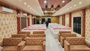 A banquet hall with rows of white tables and brown upholstered chairs, air conditioning units on the walls, and a patterned wallpaper design. - Aman Hotel (Pindra)
