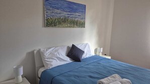 Ein ordentlich bezogenes Bett mit blauer Bettdecke und Kissen, ein weißer Nachttisch mit Lampe und ein Landschaftsbild an der Wand.