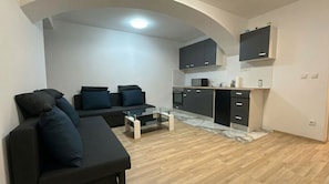 Ein kompakter Wohnbereich mit einer Couch, einem Glastisch, einer Küche mit dunklen Schränken und einem Holzboden.
