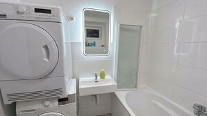 Ein modernes Badezimmer mit gestapelter Waschmaschine und Trockner, einem Waschtisch inklusive Spiegel, einer Badewanne und einer Dusche.