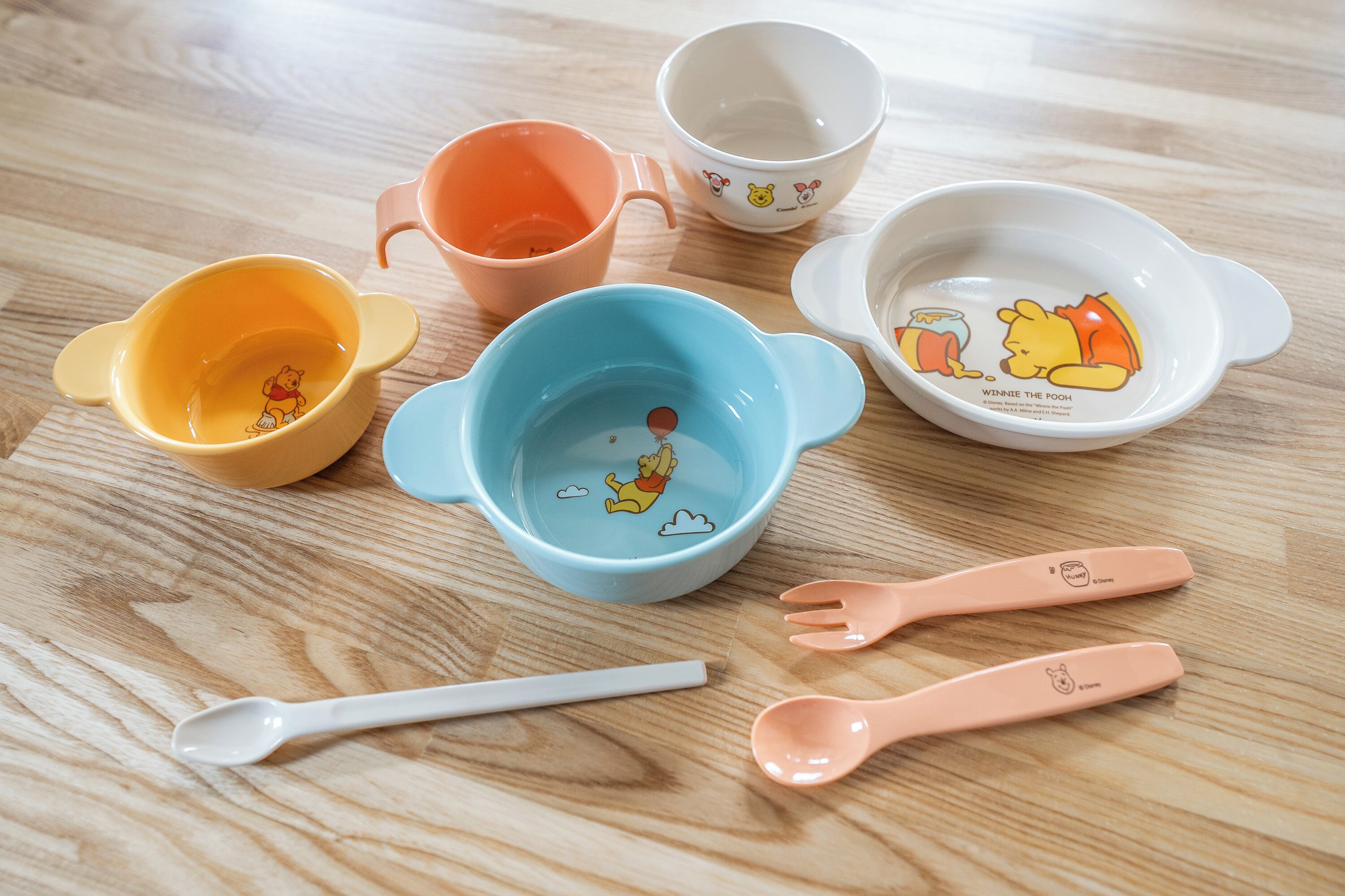 A collection of Winnie the Pooh themed bowls and spoons on a wooden table.