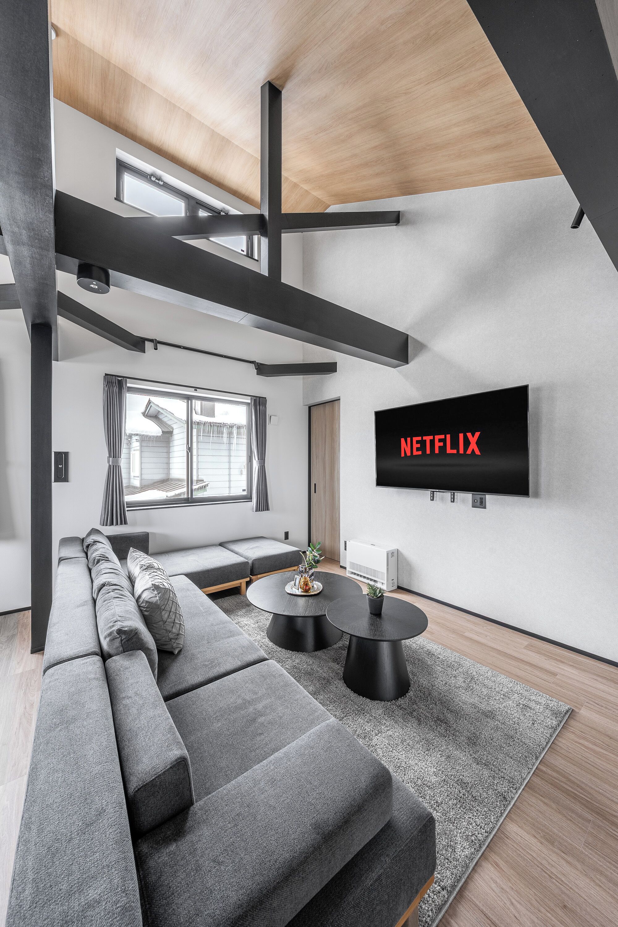 Ein modernes Wohnzimmer mit einer großen grauen Eckcouch, einem an der Wand montierten Flachbildfernseher und einem kleinen runden Tisch mit zwei schwarzen Hocker.