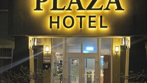 The exterior of the Plaza Hotel with illuminated signage, a double door entrance, and potted plants at the corners.