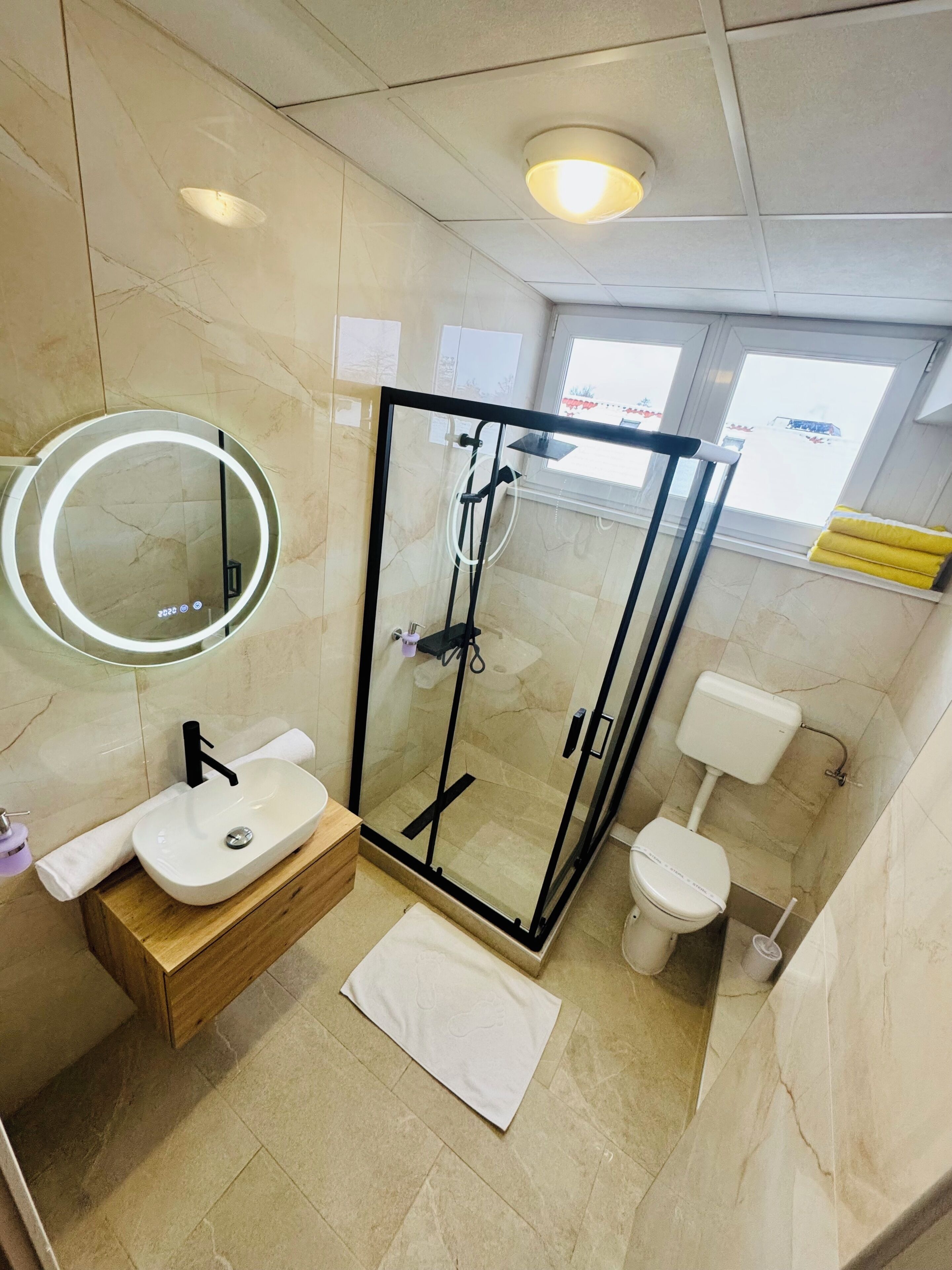 Un baño moderno con ducha de vidrio, un espejo redondo, un lavamanos con tocador de madera y un inodoro.