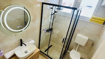 Un baño moderno con ducha de vidrio, un espejo redondo, un lavamanos con tocador de madera y un inodoro.