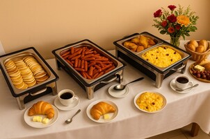 Una mesa de buffet con diversos alimentos para el desayuno, incluyendo bollería, salchichas y huevos revueltos.