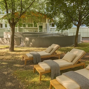 A patio area with wooden lounge chairs, a table, and a view of a building with large windows. - Pláza Hotel Gyula (Gyula)