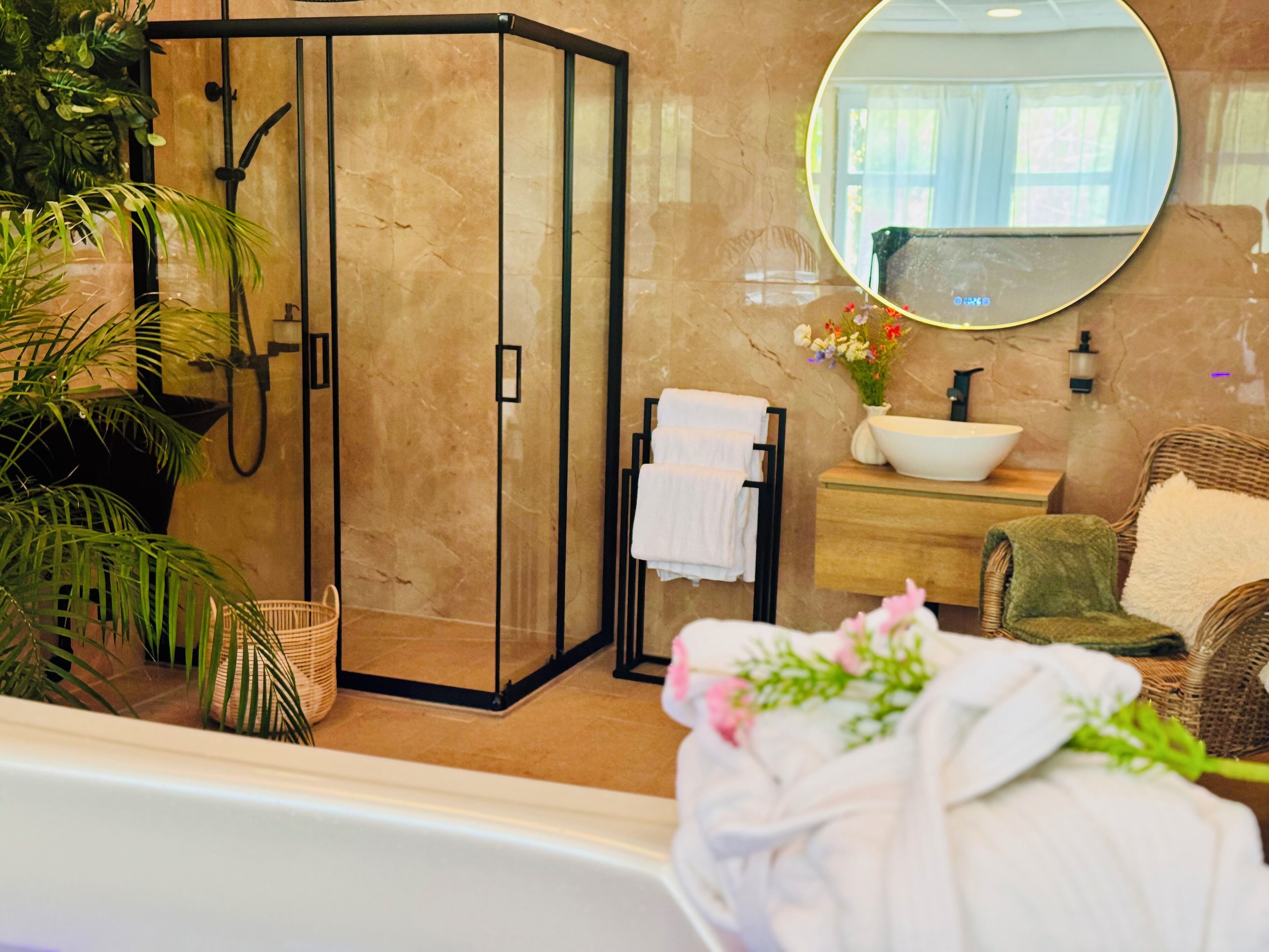 A modern bathroom with a glass-enclosed shower, a round mirror, a sink with a bowl, and a wicker chair.