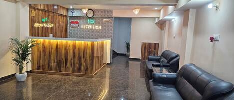 A reception area with a wooden reception desk, a clock, and a neon sign indicating 'RECEPTION'. There are black leather sofas and a potted plant.