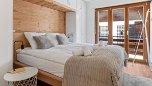 Un dormitorio moderno con cabecera de madera, una cama grande con ropa de cama blanca, una mesita redonda y vistas de la ciudad a través de una puerta de cristal.