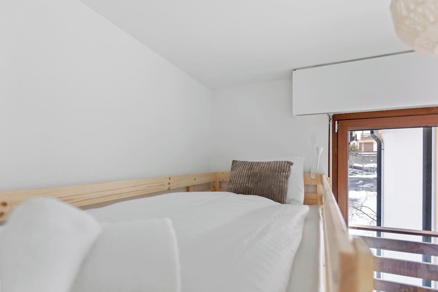A bedroom with a wooden bed, white bedding, a window with a view of buildings, and a white ceiling.