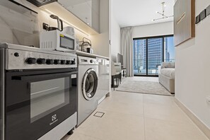 A modern kitchen with an Electrolux oven, microwave, and washing machine, a dining area with a table and chairs, and a large window with a city view. - Cozy City-View Studio w Balcony & Queen Bed (Business Bay)