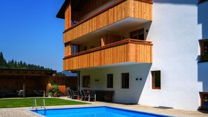 Un bâtiment moderne à deux étages, doté d’un balcon en bois, d’une piscine et d’un patio aménagé avec du mobilier d’extérieur.