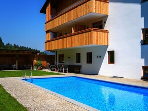 Ein modernes, zweistöckiges Gebäude mit einem hölzernen Balkon, einem Swimmingpool und einer Terrasse mit Gartenmöbeln.