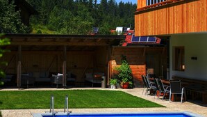 Ein Poolbereich mit überdachten Sitzgelegenheiten, einer Rasenfläche und einem Holzgebäude mit Solaranlage.