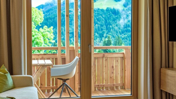 Quarto com cama, uma cadeira e varanda de madeira com vista para uma floresta e montanhas.