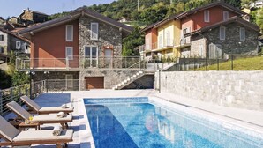 Una piscina con sillones de descanso, un edificio de piedra con balcón y una colina al fondo.