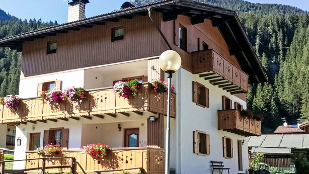 Un edificio de varios pisos con balcones decorados con macetas de flores, una chimenea visible y una vista despejada de las montañas boscosas circundantes.