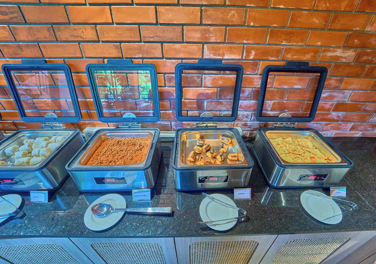 A buffet with various food items in metal containers, a brick wall in the background, and a counter with plates and utensils.