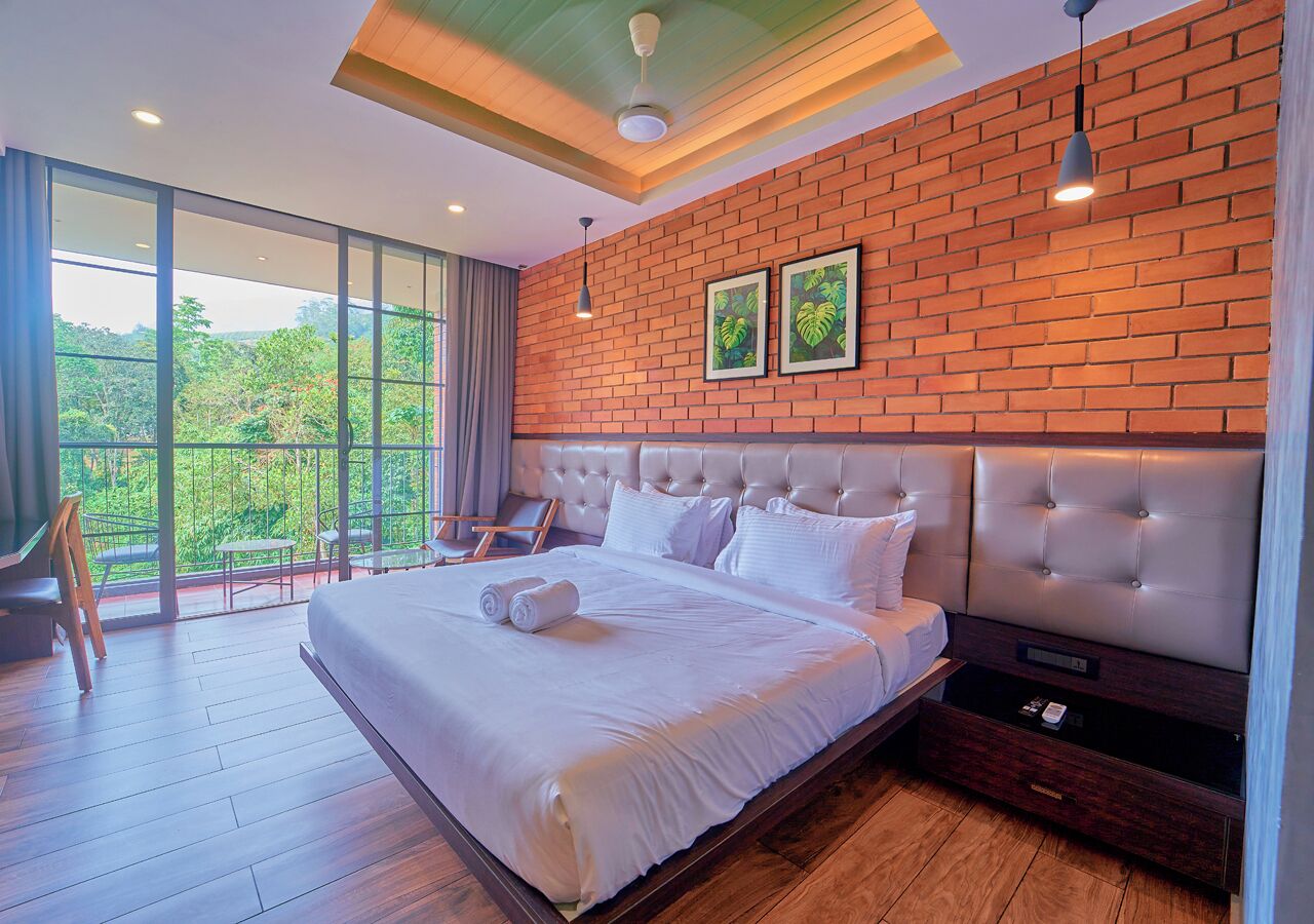 A modern hotel room with a large bed, a wooden floor, a brick accent wall, and a balcony with a view of greenery.