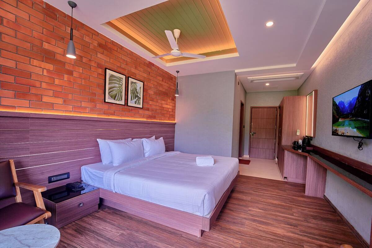 Una habitación de hotel moderna con una cama grande, piso de madera, una pared de ladrillo decorativa y un ventilador de techo, además de un televisor montado en la pared.
