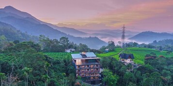 A multi-story building with a flat roof, surrounded by lush green fields and mountains in the background.