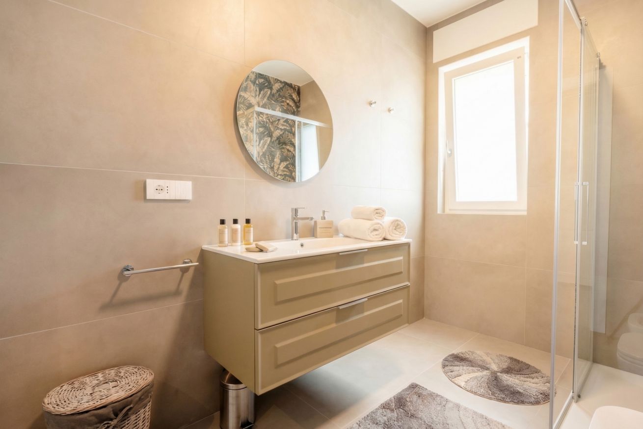 Un baño moderno con un espejo redondo, un tocador color beige, cabina de ducha de vidrio y una pequeña alfombra redonda.