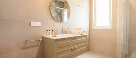 A modern bathroom with a round mirror, a beige vanity, a glass shower enclosure, and a small round rug.
