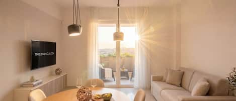 A modern living room with a flat-screen TV, a round dining table with chairs, a sofa, and a large window with natural light.