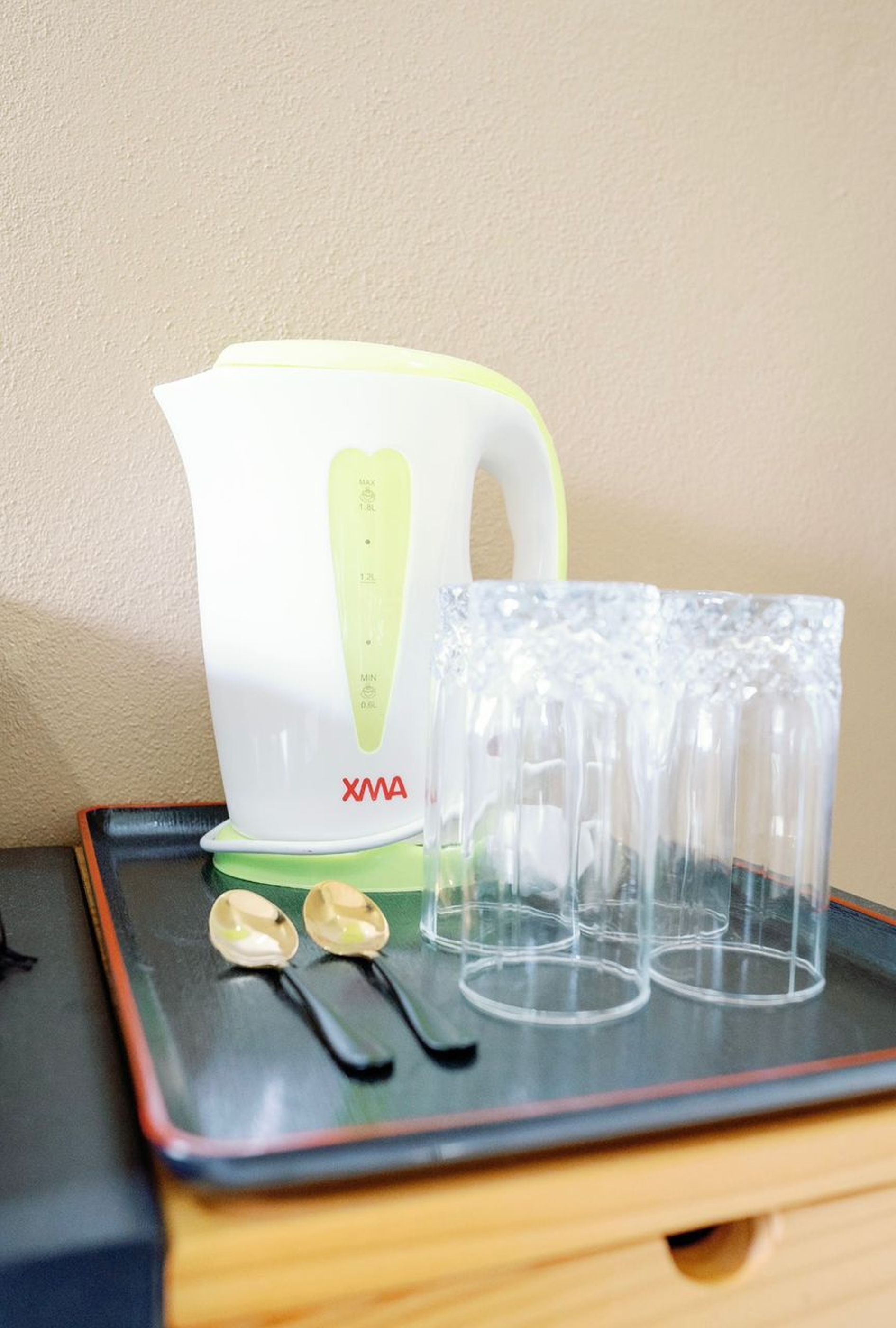 A white and green electric kettle with measurement markings, a tray with four clear glasses, and two spoons on a wooden surface.