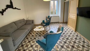 Un salon avec un canapé gris, un fauteuil bleu, une petite table sur laquelle repose un livre et une lampe suspendue jaune.