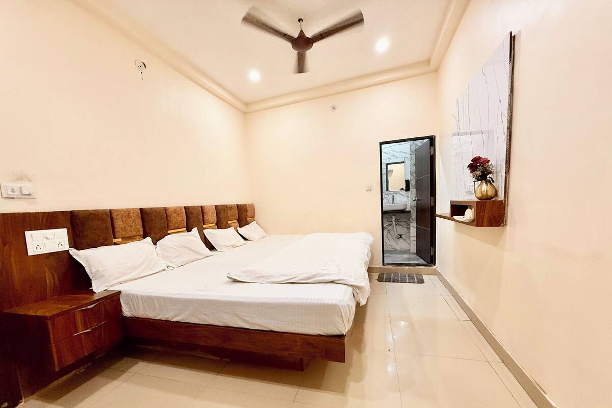 Een slaapkamer met een houten bed, nachtkastje, plafondventilator, badkamers deur en een klein plankje met een vaas.