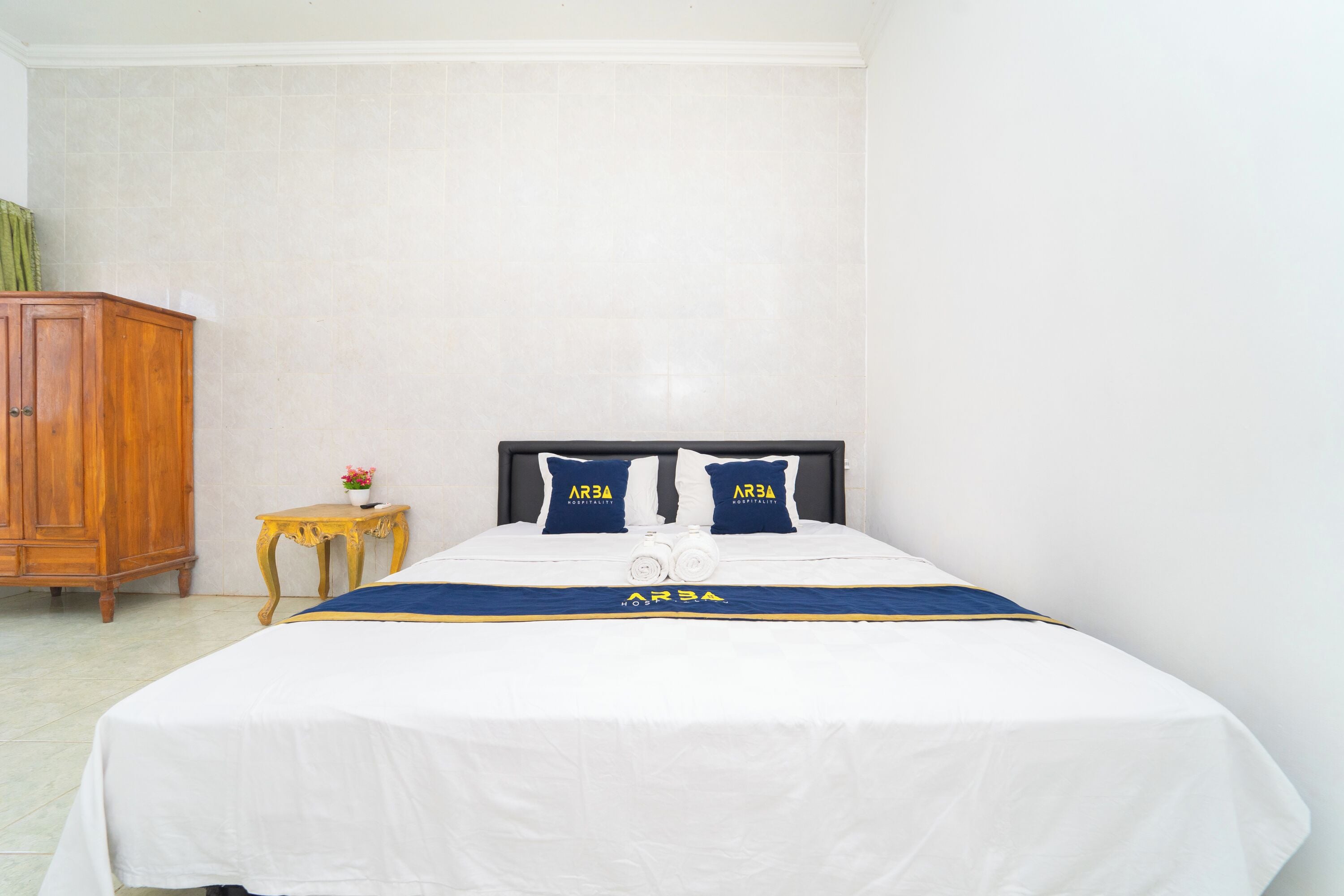 A neatly made bed with a white and blue bedspread, two matching pillows, and a small bedside table with a flower vase.